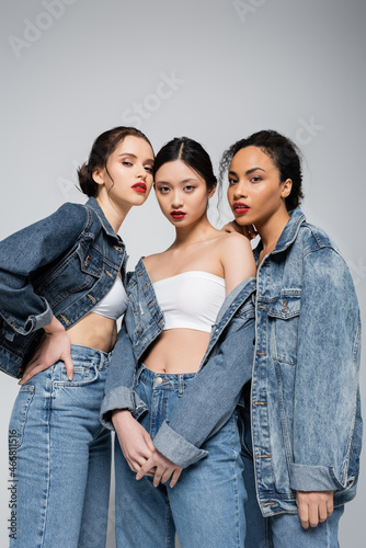 Low angle view of young interracial women in denim jackets looking at camera isolated on grey