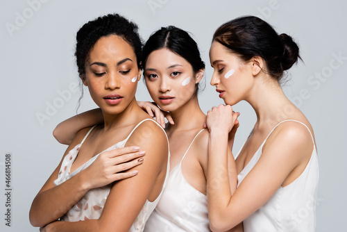 Multiethnic women with cosmetic cream on faces hugging each other isolated on grey