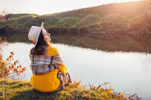 Woman relaxing by autumn lake at sunset. Stylish girl in hat sitting on bank enjoying fall landscape, breathing freely