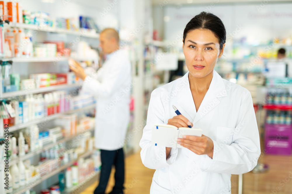 Portrait of an female pharmacist working in pharmacy during the pandemic, standing in trading floor and makes important notes