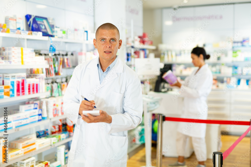 Doctor in white coat standing in salesroom with pan and recipe blanks in hands and looking in camera. His co-worker setting out drugs on shelves in background.