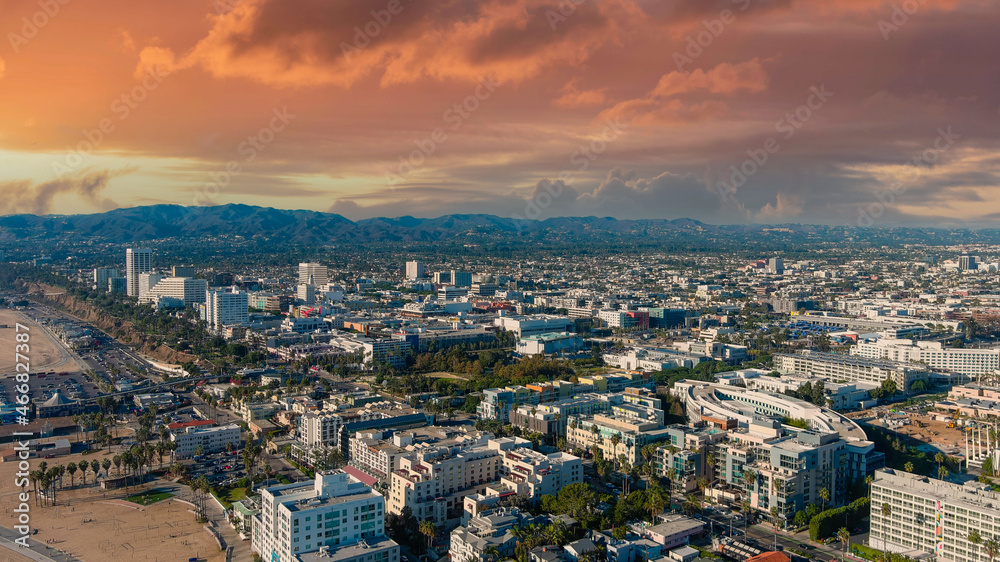 an aerial shot of the cityscape of the City of Santa Monica with vast miles of buildings and palm trees with majestic mountain ranges and powerful clouds at sunset in Santa Monica California USA - obrazy, fototapety, plakaty 