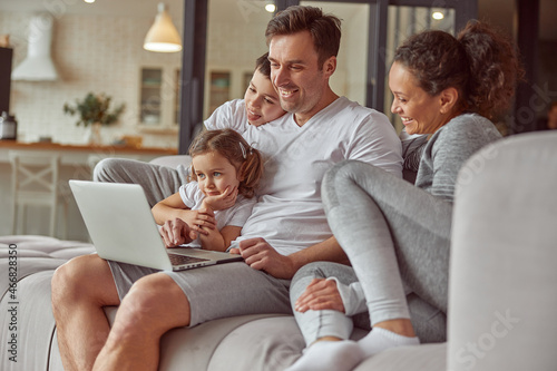 Jolly parents are cuddling with daughter and son while spending time with laptop in living room