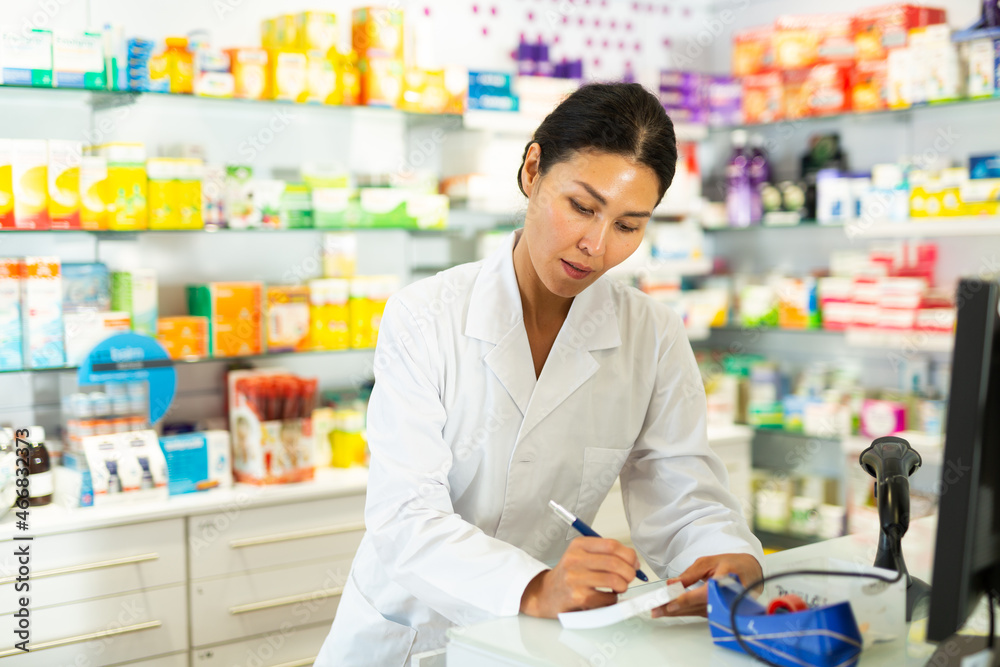 Asian woman doctor standing at counter in drugstore and writing recipe.