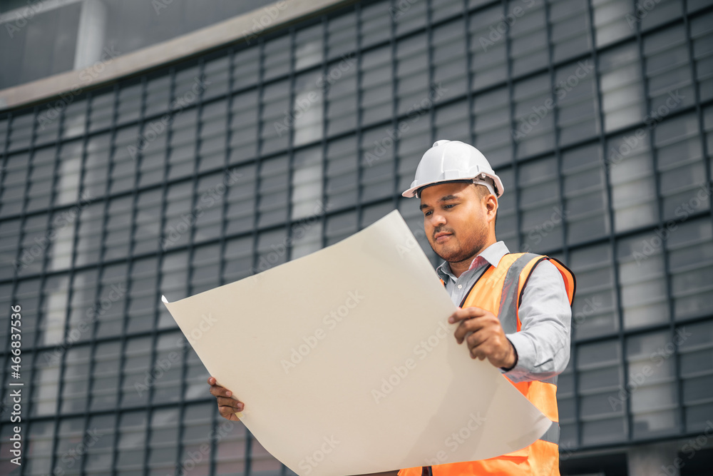 Asian engineer handsome man or architect open the paperwork blueprint and planning to building construction. Worker asian man with white safety helmet working in modern building construction site. Asian engineer handsome man or architect open the paperwork blueprint and planning to building construction. Worker asian man with white safety helmet working in modern building construction site.