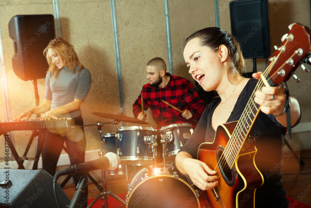Attractive cheerful smiling female soloist playing guitar and singing with her music band in sound studio