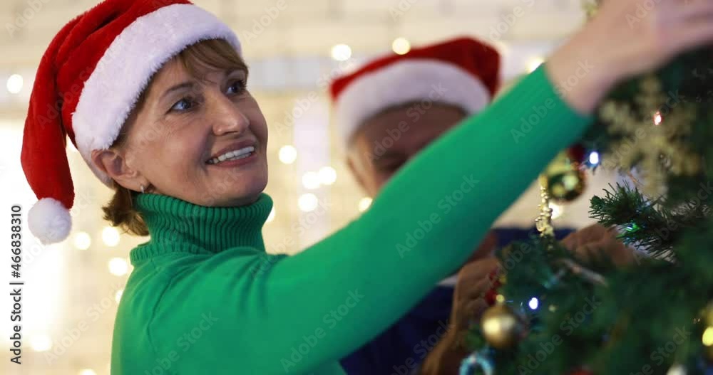Smiling elderly man and middle aged female in Santa hats decorating coniferous holiday tree and celebrating Christmas together at home