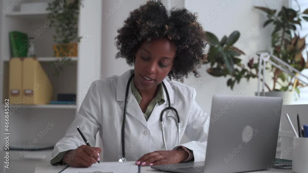 Doctor at distance appointment via computer. Afro-American woman in white robe fills spbas in medical card talking to patient via laptop at workplace in clinic Doctor at distance appointment via computer. Afro-American woman in white robe fills spbas in medical card talking to patient via laptop at workplace in clinic