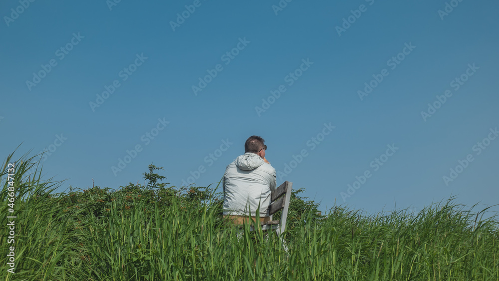 On a green meadow, among lush grass, there is a bench. A man in a windbreaker is sitting against a clear blue sky. View from the back. Kamchatka On a green meadow, among lush grass, there is a bench. A man in a windbreaker is sitting against a clear blue sky. View from the back. Kamchatka