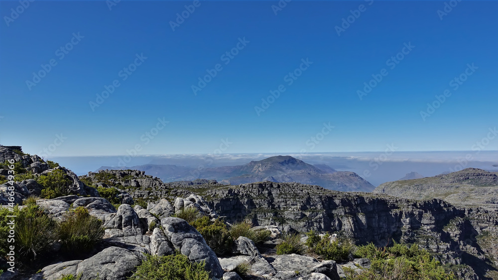 At the top of Table Mountain in Cape Town there is scant vegetation, gray boulders. Clear blue sky. A sunny summer day. South Africa