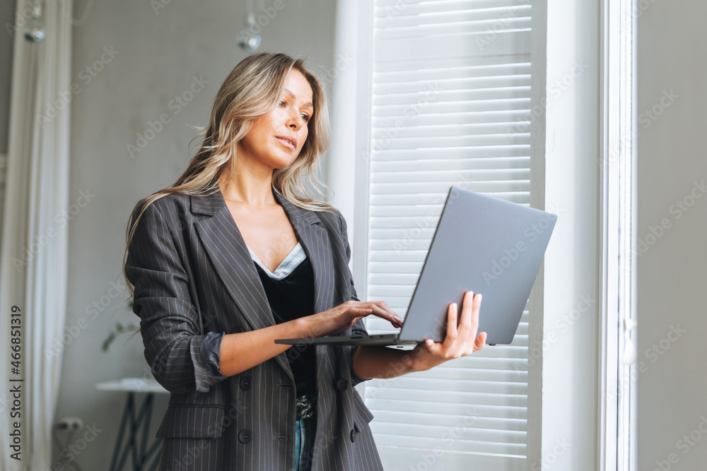 Young smiling blonde woman with long hair in stylish grey suit working at laptop in the bright modern office Young smiling blonde woman with long hair in stylish grey suit working at laptop in the bright modern office