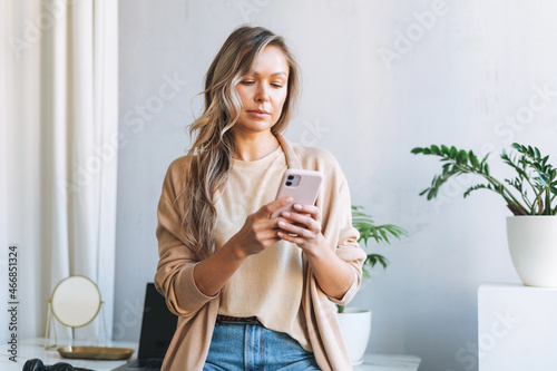 Young smiling blonde woman with long hair in stylish cardigan working at laptop in the bright modern office, woman send message by smartphone