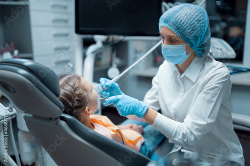 Side view of dentist in medical uniform who treats teeth of little girl patient in dental office