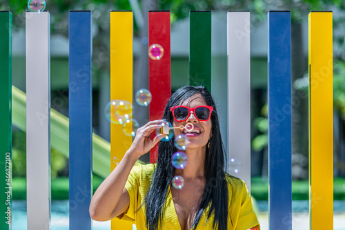 Latina woman smiling and blowing bubbles soap in a yellow dress against a background of a pool and colorful bars on a sunny day
