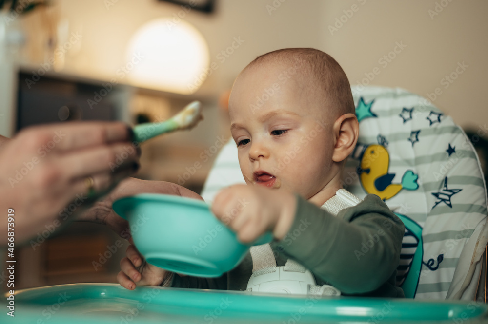 Baby eating with a face stained in food while mother feeds him with a spoon Baby eating with a face stained in food while mother feeds him with a spoon