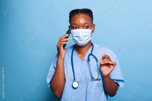 African american practitioner assistant with protective face mask against coroanvirus discussing with collegue at phone explaining sickness expertise while working in studio. Medicine concept photo