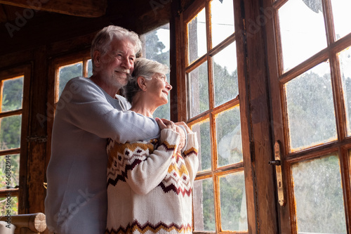 Elderly age relationship and love concept with couple of mature man and woman hugging and looking outside the windows at home with happiness and joy together photo