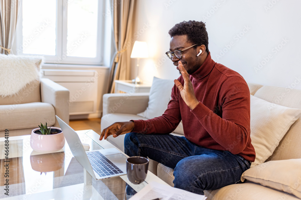 Cute handsome African American guy social distancing at home sitting in living room using laptop looking at screen and waving hand saying Hi while chatting online with friends, having video call