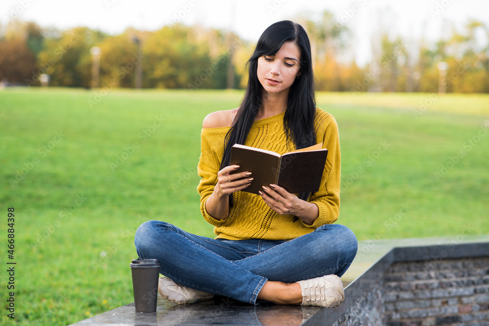Obraz premium A beautiful and attractive Caucasian brunette girl in a yellow sweater is reading a book while sitting in the park.