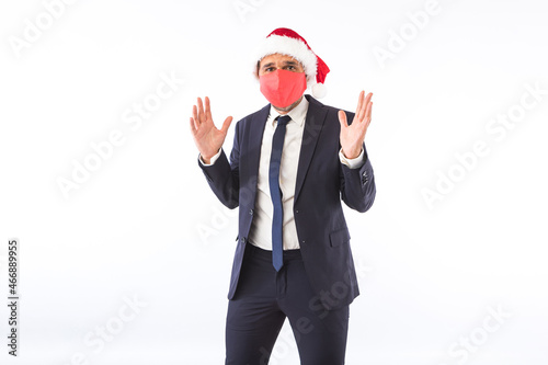 Businessman dressed in suit, tie, Santa Claus Christmas hat and red mask, raising hands having fun, on white background. Christmas celebration concept. © Davidbenito