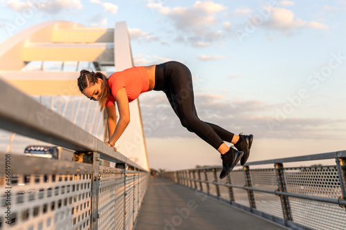 Young happy focused fitness girl in black yoga pants and orange short shirt jumps from bridge fence on footpath. Front view.