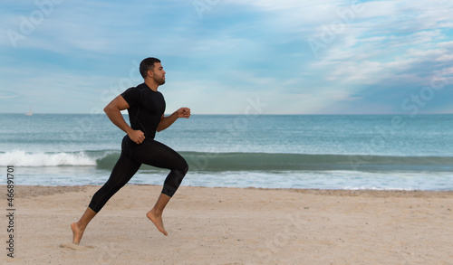 hombre moreno y fuerte de cuerpo atlético indio o pakistaní practica deporte y corre con ropa ajustada negra en un playa de arena con un fondo azul 