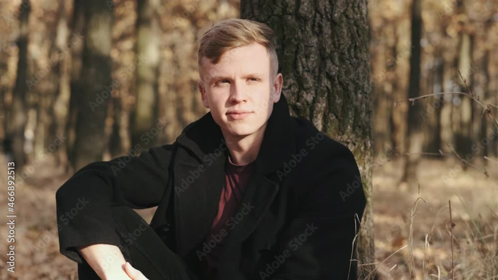 Portrait of handsome man in black coat sitting near the tree in autumn forest on sunny day Portrait of handsome man in black coat sitting near the tree in autumn forest on sunny day