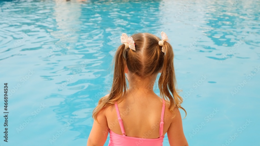 joyful little girl 4-5 years old, with ponytails, dressed in a pink swimsuit, looks at the blue water in the pool or in the sea on summer vacation, back view joyful little girl 4-5 years old, with ponytails, dressed in a pink swimsuit, looks at the blue water in the pool or in the sea on summer vacation, back view