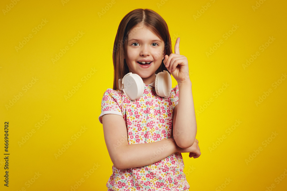 Optimistic kid in summer clothes and white headphones on neck smiling for camera and pointing up while having great idea against yellow background Optimistic kid in summer clothes and white headphones on neck smiling for camera and pointing up while having great idea against yellow background