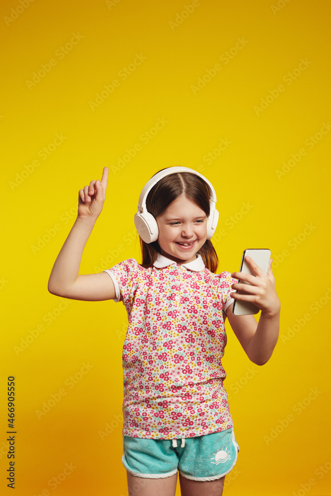 Vertical photo of little kid with white headphones listen music pointing index finger up while looking at smartphone isolated on yellow background Vertical photo of little kid with white headphones listen music pointing index finger up while looking at smartphone isolated on yellow background