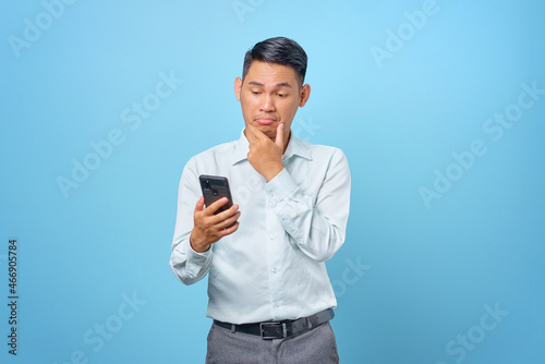 Pensive young handsome businessman using smartphone on blue background