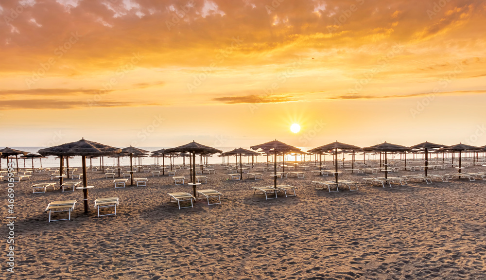 scenic view at nice sunset or sunrise beach with rows of umbrellas and chaise lounges with blue sea and amazing cloudy sky with sun glow on the background