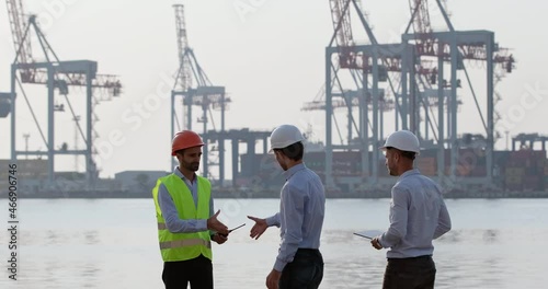 Men engineers and port worker wearing protective helmets discuss work issues concerning dockside cranes loading process on foggy morning