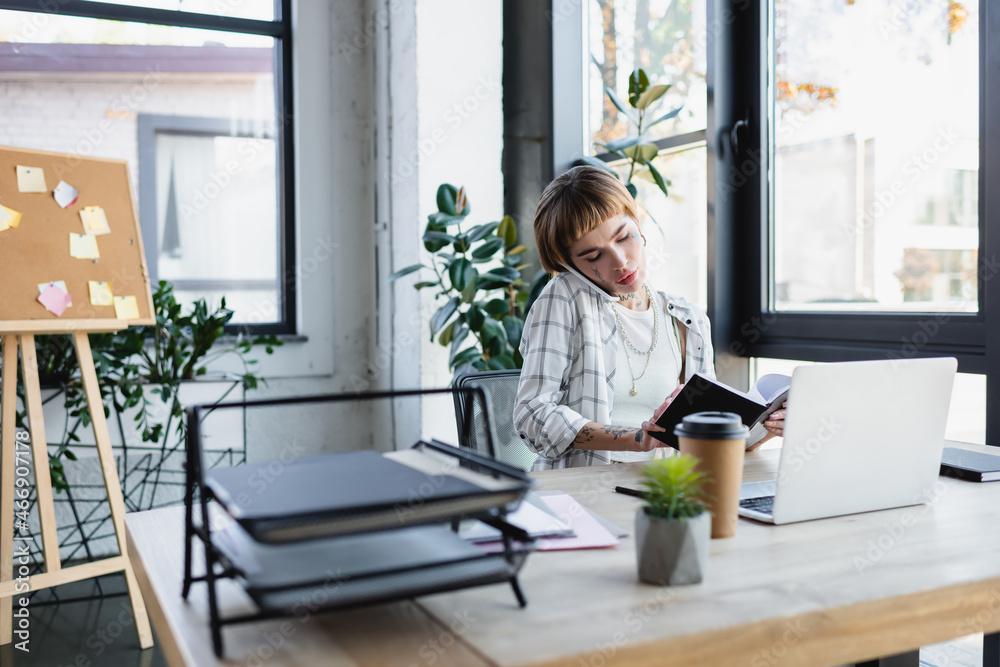 young tattooed woman with notebook calling on smartphone near laptop in office young tattooed woman with notebook calling on smartphone near laptop in office