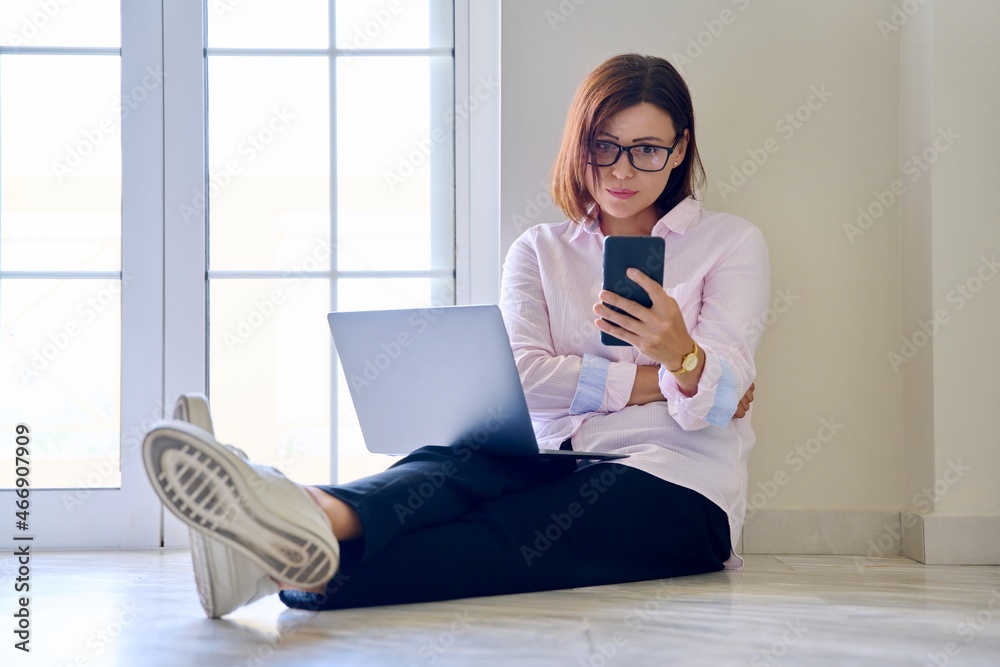 Business woman sitting on the floor with a laptop looking at smartphone Business woman sitting on the floor with a laptop looking at smartphone