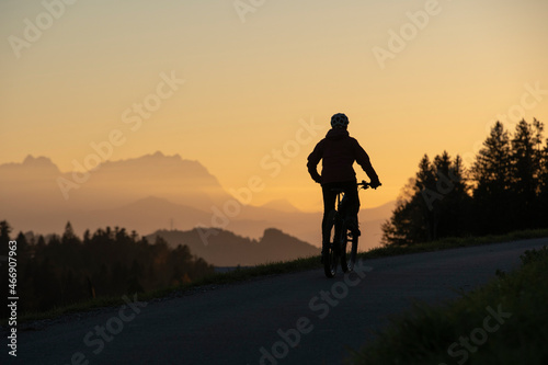 Fototapeta Naklejka Na Ścianę i Meble -  active woman riding her electric mountain bike at sunset in front of the awesome silhouette of Mount Saentis, Appenzell switzerland