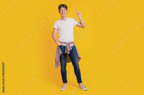 Full length photo of young cheerful guy standing hand in pocket show peace cool v-sign isolated over yellow color background