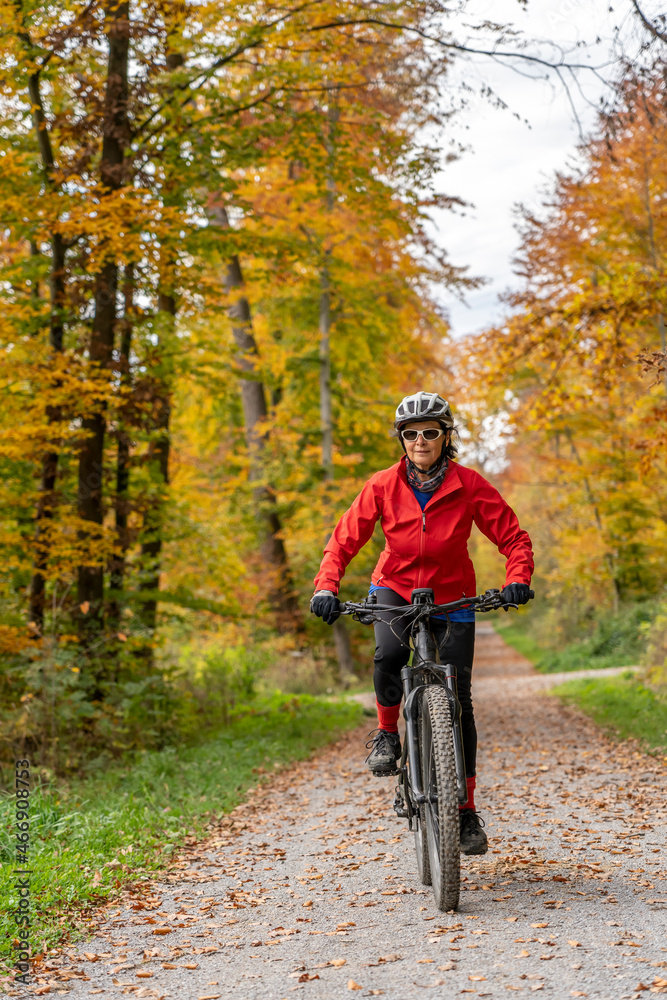 pretty senior woman ridin her electric bicycle in a colorful autumn forest with golden foliage
