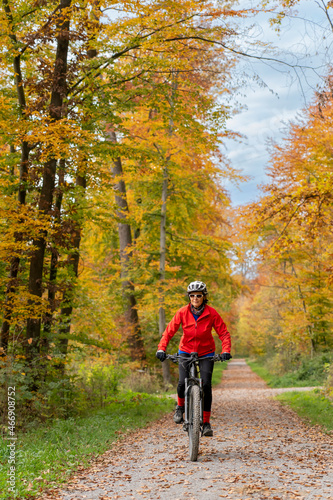 pretty senior woman ridin her electric bicycle in a colorful autumn forest with golden foliage 