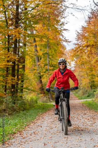 pretty senior woman ridin her electric bicycle in a colorful autumn forest with golden foliage
