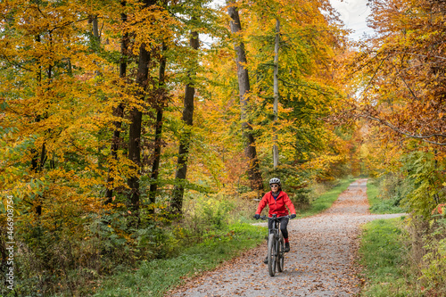 pretty senior woman ridin her electric bicycle in a colorful autumn forest with golden foliage 