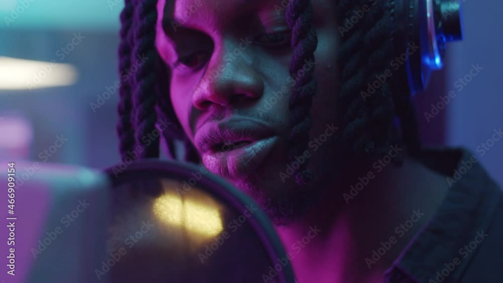 Close up shot of African American man with dreadlocks singing in microphone in recording studio with neon purple light