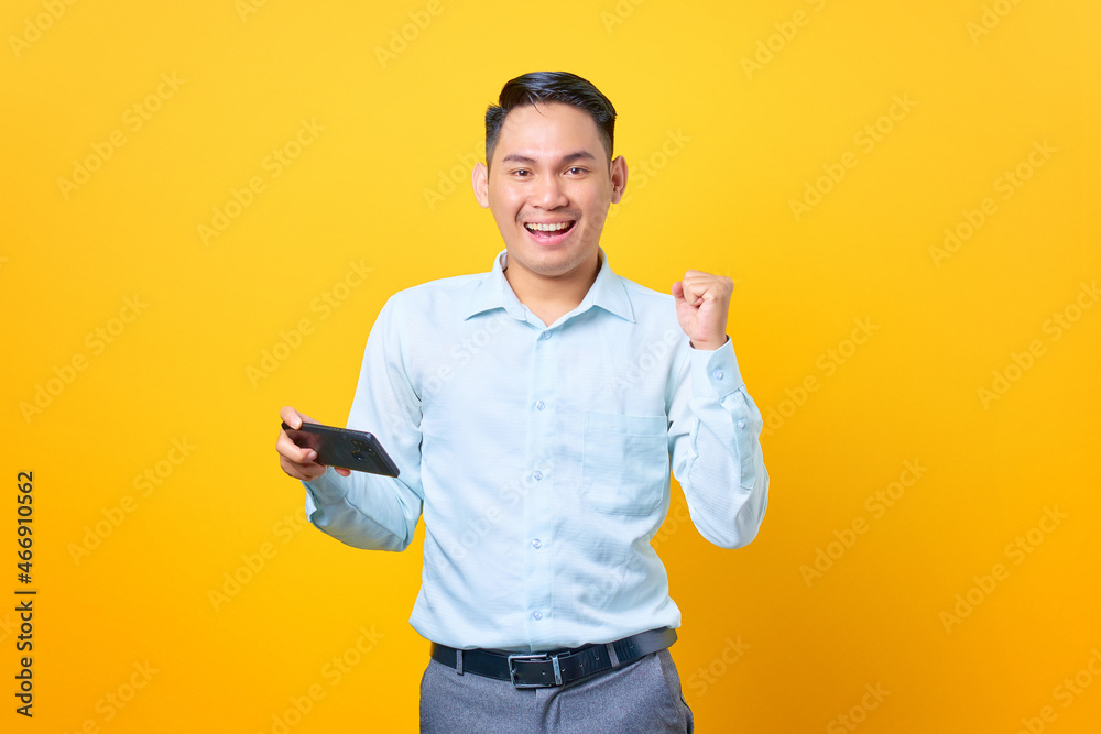 Excited young handsome businessman using smartphone and doing victory gesture on yellow background Excited young handsome businessman using smartphone and doing victory gesture on yellow background