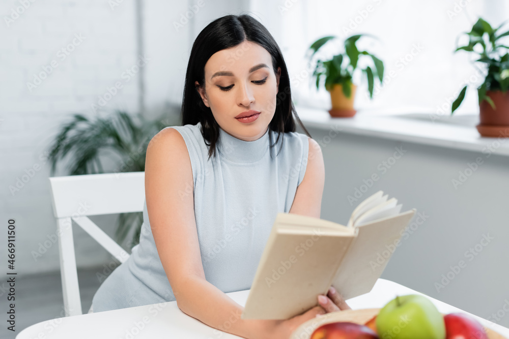 young woman reading book near blurred apples on kitchen table young woman reading book near blurred apples on kitchen table