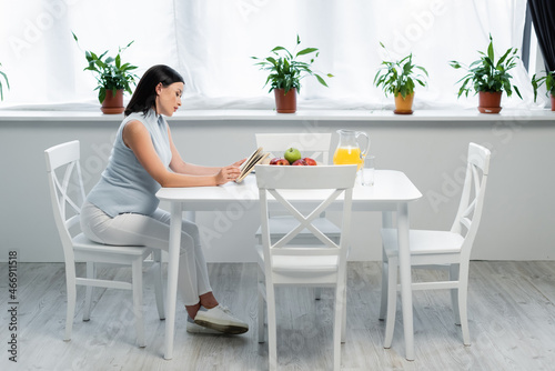 side view of pregnant woman reading book near orange juice and fresh apples on table in kitchen