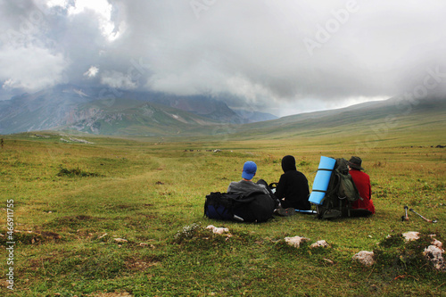 a group of tourists with backpacks sit and look at the green valley and mountains in heavy clouds