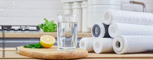 Glass of filtered clean water with reverse osmosis filter, lemons and cartridges on table in kitchen. Concept Household filtration or purification system.