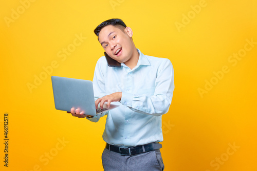 Young handsome businessman talking on a smartphone while holding a laptop on yellow background