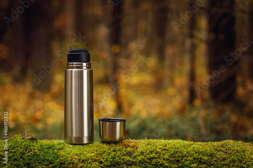 Steel thermos with delicious hot tea on an old mossy tree in focus. The background of the autumn forest is blurred.