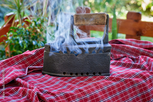 Cast-iron ancient iron with a wooden handle, with a compartment for coal, stands on clothes for ironing. Hot, vintage old iron with coal, selective focus.